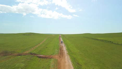 Aerial-Drone-View-Of-A-Vehicle-Driving-Across-The-Isolated-Dirt-Road-Between-Green-Grassland