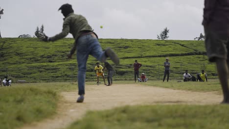 Partido-De-Críquet-En-Un-Campo-De-Tierra-Rural-Con-Un-Exuberante-Telón-De-Fondo-De-Plantaciones-De-Té-Verde-En-Sri-Lanka