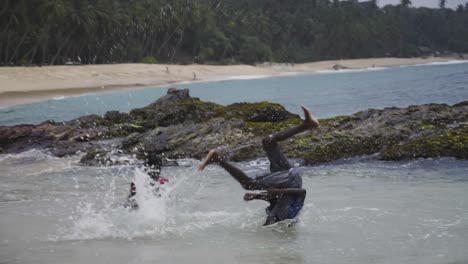 Two-boys-playing-in-the-waves-near-rocky-shoreline-on-a-tropical-beach