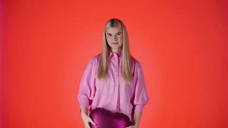 Pretty-Blonde-Woman-Throwing-Purple-Heart-Shaped-Balloons-Up-In-Air-Against-Red-Background,-Studio-Shot