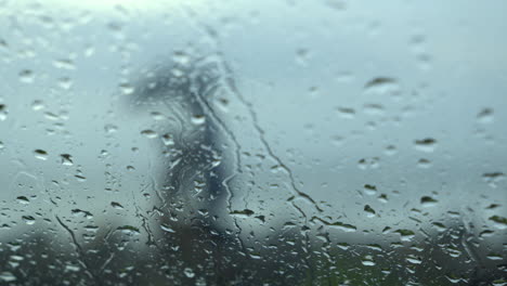 Gotas-De-Lluvia-Goteando-Sobre-El-Cristal-Húmedo-Y-Transparente-De-La-Ventanilla-Del-Coche-Durante-Las-Fuertes-Lluvias-Contra-La-Vista-De-La-Playa,-La-Gente-Disfruta-Del-Día-Lluvioso-En-La-Temporada-De-Monzones