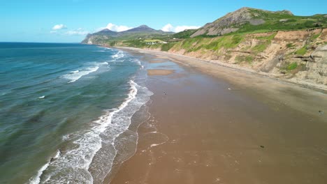 Nefyn-beach-fast-drone-high-level-sweep-over-waves-breaking,-beautiful-North-Wales-coastline,-UK