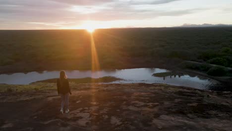 Mujer-Caminando-Hacia-El-Atardecer-Sobre-Un-Terreno-Rocoso-Con-Una-Vista-Panorámica-De-La-Sabana-Y-El-Río.