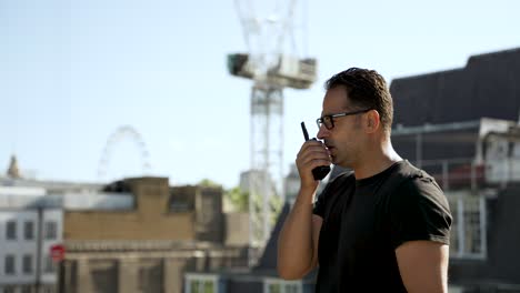 Man-in-a-black-t-shirt-using-walkie-talkie-on-a-rooftop-with-a-clear-view-of-an-urban-skyline-featuring-buildings-and-greenery,-showcasing-communication-in-a-city-setting