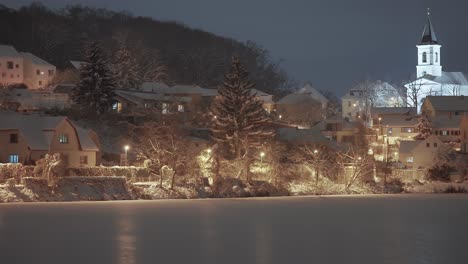 Una-Serena-Noche-De-Invierno-Con-Casas-Cubiertas-De-Nieve-Y-Una-Iglesia,-Iluminada-Por-Farolas