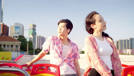 Modern-couple-wearing-vibrant-colorful-shirt-listening-to-music-with-wireless-earphones-standing-on-open-air-bus-traveling-around-modern-skyline-city