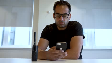 Male-wearing-glasses-is-seated-at-a-white-table,-focused-on-using-his-smartphone