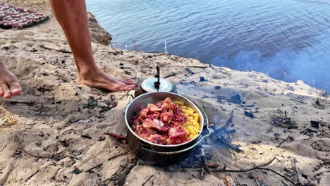 Food-cooking-in-a-pot-on-fire-on-the-beach-in-the-sand,-a-man-adds-sauce,-slow-motion