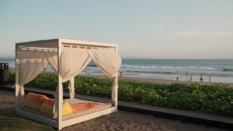 W-Hotel-Luxury-Outdoor-Patio-Sunbed-Gazebo-With-Curtain,-Cushions,-Pillows,-and-Canopy-in-Outdoor-Beachfront-Lounge,-People-Walking-by-the-Sea-in-Slow-Motion-At-Sunset-Beach,-Seminyak,-Bali-Indonesia