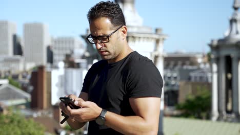 Athletic-European-man-with-glasses-and-walkie-talkie,-using-a-smartphone-outdoors-against-an-urban-backdrop