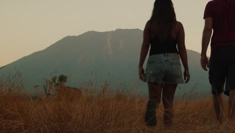 Couple-walking-through-Tianyar-savanna-in-Bali,-Indonesia,-with-active-Vulcano-in-the-background