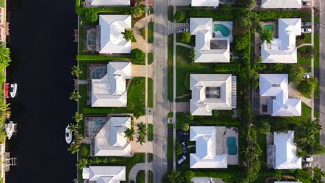 Aerial-view-of-a-waterfront-neighborhood-with-white-roofed-houses,-green-lawns,-and-swimming-pools,-arranged-neatly-along-a-canal-with-docked-boats