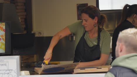 in-a-pizzeria-trailer,-a-pretty-brunette-lady-in-a-black-apron-cutting-up-a-pizza-and-closing-her-box-as-she-greets-a-customer,-another-employee-behind-her,-stacks-of-boxes-in-the-background