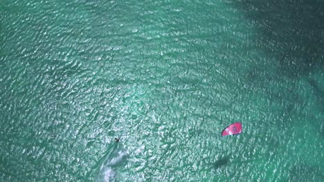 Kitesurfer-gliding-over-clear-turquoise-water-with-a-red-kite,-aerial-view