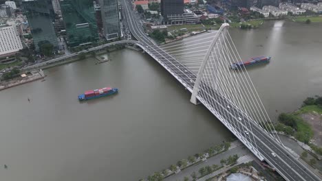 Container-boats-on-Saigon-River,-Ho-Chi-Minh-City,-with-high-aerial-view-of-Ba-Son-Bridge-as-boat-pass-under