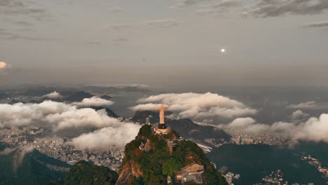 Drohnenaufnahmen-Der-Ikonischen-Christusstatue,-Beleuchtet-Vom-Vollmond,-Mit-Blick-Auf-Die-Pulsierende-Stadtlandschaft-Von-Rio-De-Janeiro-Bei-Sonnenuntergang