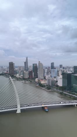 Container-boats-on-Saigon-River,-Ho-Chi-Minh-City,-with-high-aerial-view-of-Ba-Son-Bridge-with-City-Skyline-in-vertical-format-as-boat-passes-under