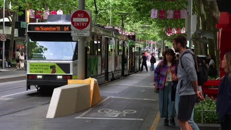 Tram-arrived-at-the-stop-with-commuters-disembarking-and-boarding-the-tram-during-rush-hour-in-Melbourne-city's-bustling-Central-Business-District,-pedestrians-crossing-tree-lined-Swanston-street