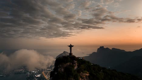 Erleben-Sie-Die-Atemberaubende-Luftaufnahme-Der-Christusstatue-In-Rio-De-Janeiro,-Beleuchtet-Von-Den-Goldenen-Farbtönen-Eines-Atemberaubenden-Sonnenuntergangs,-Mit-Der-Lebendigen-Stadtlandschaft-Und-Dem-Heiteren-Himmel