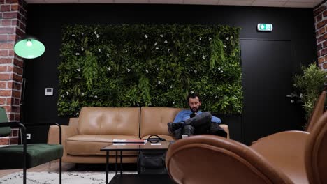 a-dark-haired,-bearded-man-sitting-alone-on-a-beige-leather-sofa,-legs-crossed,-in-an-open-space,-a-green-wall-behind-him