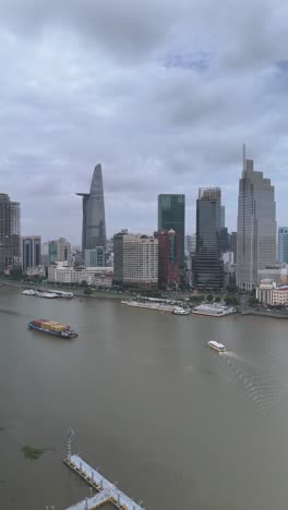 Container-boats-and-ferry-on-Saigon-River,-Ho-Chi-Minh-City,-with-aerial-panning-view-of-city-skyline-in-vertical-format-as-boats-pass-along-river