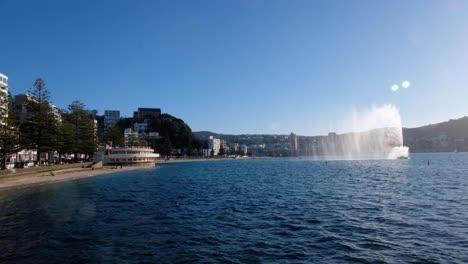 Scenic-view-overlooking-harbour-with-water-fountain,-white-sandy-beach-and-people-walking-along-waterfront-of-Oriental-Parade-in-Wellington,-New-Zealand-Aotearoa