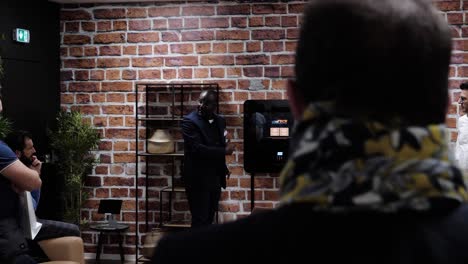 in-a-shared-open-space-in-a-company,-a-black-man-presents-and-explains-to-his-attentive-team-how-the-new-device-works,-red-brick-wall-at-the-back,-black-wall-on-the-sides,-clean-industrial-style-decor