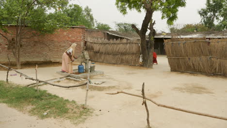 Mujer-Bombeando-Agua-En-Un-Pozo-Mientras-Otra-Mujer-Barre-En-La-India-Rural