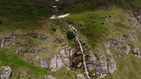 Tiered-mountain-waterfall-cascades-down-rugged-cliffs-on-a-sunny-day,-rising-drone-shot