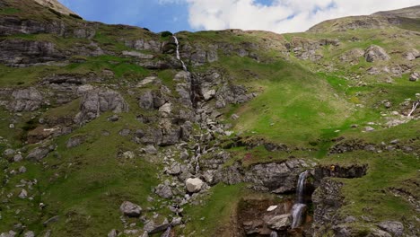 Tiered-mountain-waterfalls-cascades-down-rugged-cliffs-and-pieces-of-ice-on-a-sunny-day,-tourist-taking-photos,-drone-tracking-shot