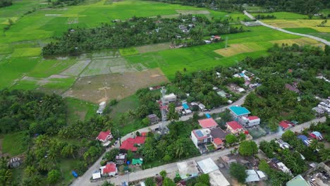 Toma-Aérea-Inclinada-Hacia-Abajo-De-Una-Torre-De-Telecomunicaciones-En-Un-Pueblo-Rural-Filipino-Con-Densas-Selvas-Y-Arrozales-En-Catanduanes