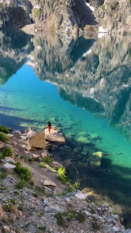 Vertical-4k,-Woman-in-Bikini-on-Rock-by-Beautiful-Glacial-Lake-With-Sky-and-Hills-Mirror-Reflection