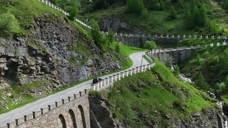 Aerial-View-Of-A-picturesque-mountain-road-winds-through-lush-green-terrain,-offering-a-stunning-view-of-the-natural-landscape-with-a-car-driving-along-the-scenic-route