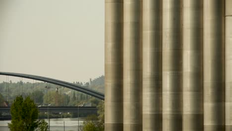 Große-Industriesäulen-Mit-Fernblick-Auf-Eine-Brücke-Und-Einen-Fluss-Bei-Sanftem-Tageslicht,-Die-Eine-Urbane-Szene-Am-Flussufer-Einfangen