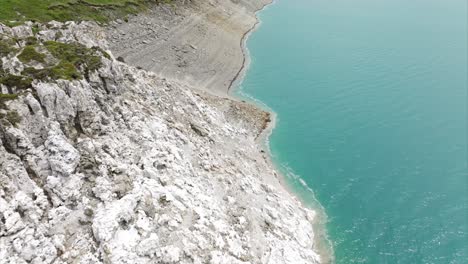 Aerial-view-looking-down-at-a-rocky-cliff-coastline-merging-into-a-serene-turquoise-sea,-showcasing-the-natural-beauty-and-rugged-terrain-of-coastal-landscapes