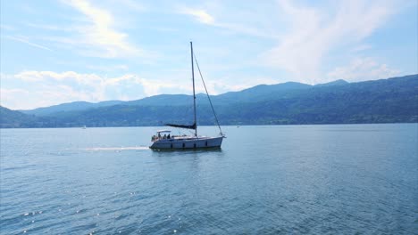 Vista-Aérea-Del-Velero-Navega-Pacíficamente-En-El-Lago-Maggiore,-Rodeado-De-Majestuosas-Montañas-En-Un-Día-Soleado,-Ofreciendo-Una-Vista-Pintoresca-De-La-Naturaleza-Para-El-Ocio-Y-La-Relajación