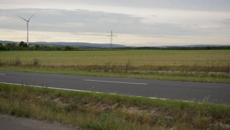 Blurred-car-speeding-by-with-wind-turbine-and-open-field-in-background,-shot-at-dusk