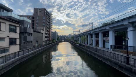 Sunset-over-a-canal-with-reflections,-cityscape,-and-train-tracks-on-the-side