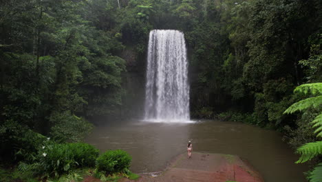 Toma-De-Drone-De-Una-Joven-Caminando-Bajo-Una-Pintoresca-Cascada-En-La-Selva-Verde,-Millaa-Milla-Falls,-Australia