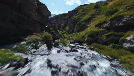 FPV-Aéreo-Volando-Por-Un-Arroyo-De-Montaña-Que-Fluye-A-Través-De-Exuberantes-Colinas-Verdes,-Dirigiéndose-Hacia-Un-Tranquilo-Lago-Turquesa,-Con-Pintorescas-Montañas-Al-Fondo