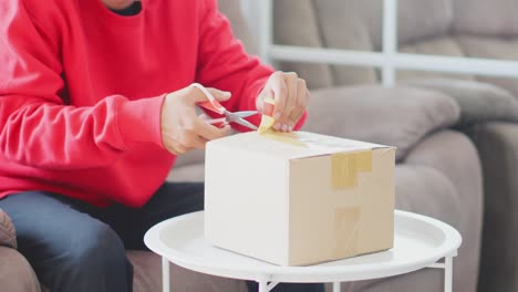 Young-Man-Sitting-On-Sofa,-Using-A-Scissor-To-Cut-Open-A-Duct-Tape-On-Cardboard-Box-At-Home