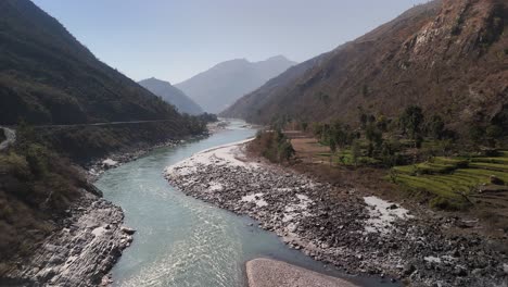 Un-Puente-Colgante-Se-Extiende-Sobre-El-Río-Rodeado-De-Campos-En-Terrazas-Y-Montañas-Con-Vegetación-En-El-Remoto-Campo-De-Nepal.