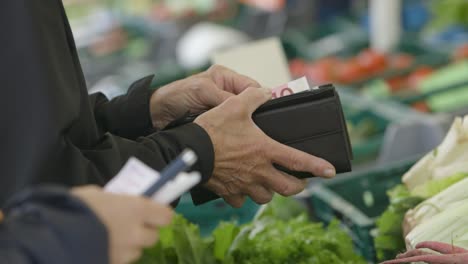 Close-up-of-a-man-paying-cash-for-groceries-at-a-market,-vivid-vegetables-in-background