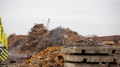 Yellow-locomotive-at-a-scrapyard-with-piles-of-metal-debris-under-cloudy-skies,-close-up