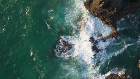 Top-down-view-of-ocean-waves-smashing-into-Cornwall-sea-cliffs,-UK