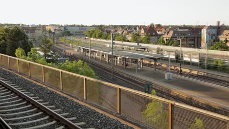 Railroad-tracks-in-foreground-leading-towards-a-train-station-at-golden-hour,-with-passengers-waiting-and-a-cityscape-background