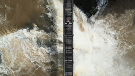 Top-Down-Aerial-View-of-Woman-Walking-on-Footbridge-Above-Powerful-Loud-Waterfall,-High-Angle-Drone-Shot