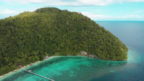 Aerial-view-of-the-lush,-forested-hills-of-Kri-Island-in-Raja-Ampat,-Indonesia,-with-turquoise-waters-and-a-long-pier-extending-into-the-sea