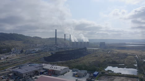 Landscape-view-with-a-coal-fired-power-station-with-its-chimneys-and-funnels-releasing-white-smoke-into-the-air-on-a-sunny-and-cloudy-day