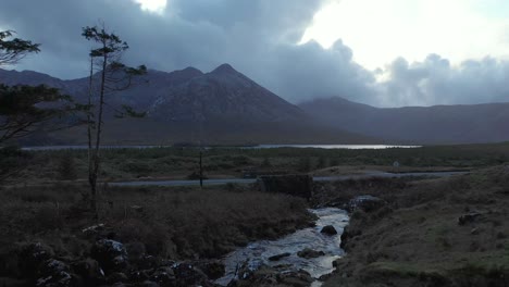 Aerial:-Car-crossing-on-empty-road-next-to-creek-and-mountain-in-irish-landscape